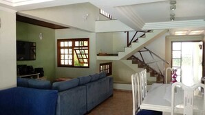 A spacious living area with a blue sofa, a dining table with chairs, and a staircase leading to an upper level. - Cond Hanga Roa - Bertioga Riviera (Bertioga)