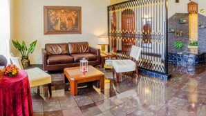 A spacious living area with a brown leather sofa, a wooden coffee table, and a white armchair. A chandelier hangs from the ceiling, and a decorative painting is on the wall. - Hotel Carmel (Lima)