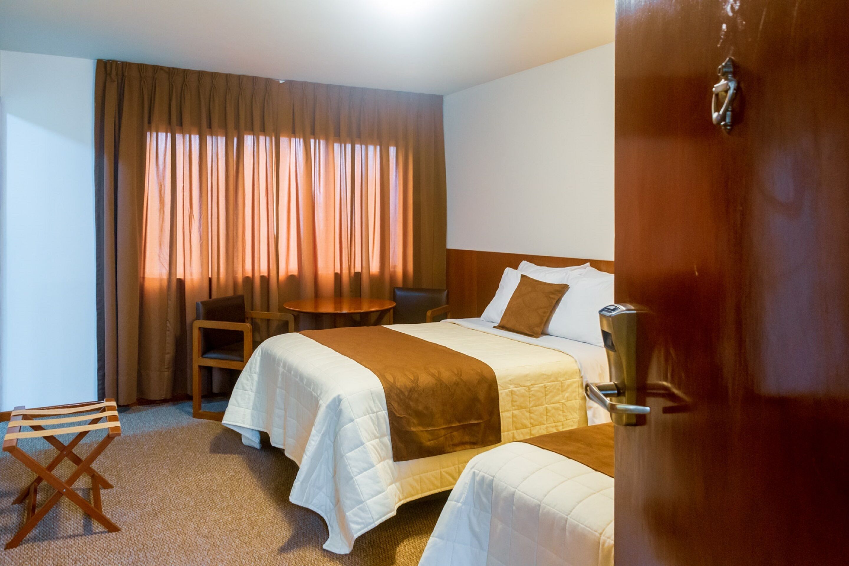 A hotel room with two beds, a small table, a chair, and a window with curtains.