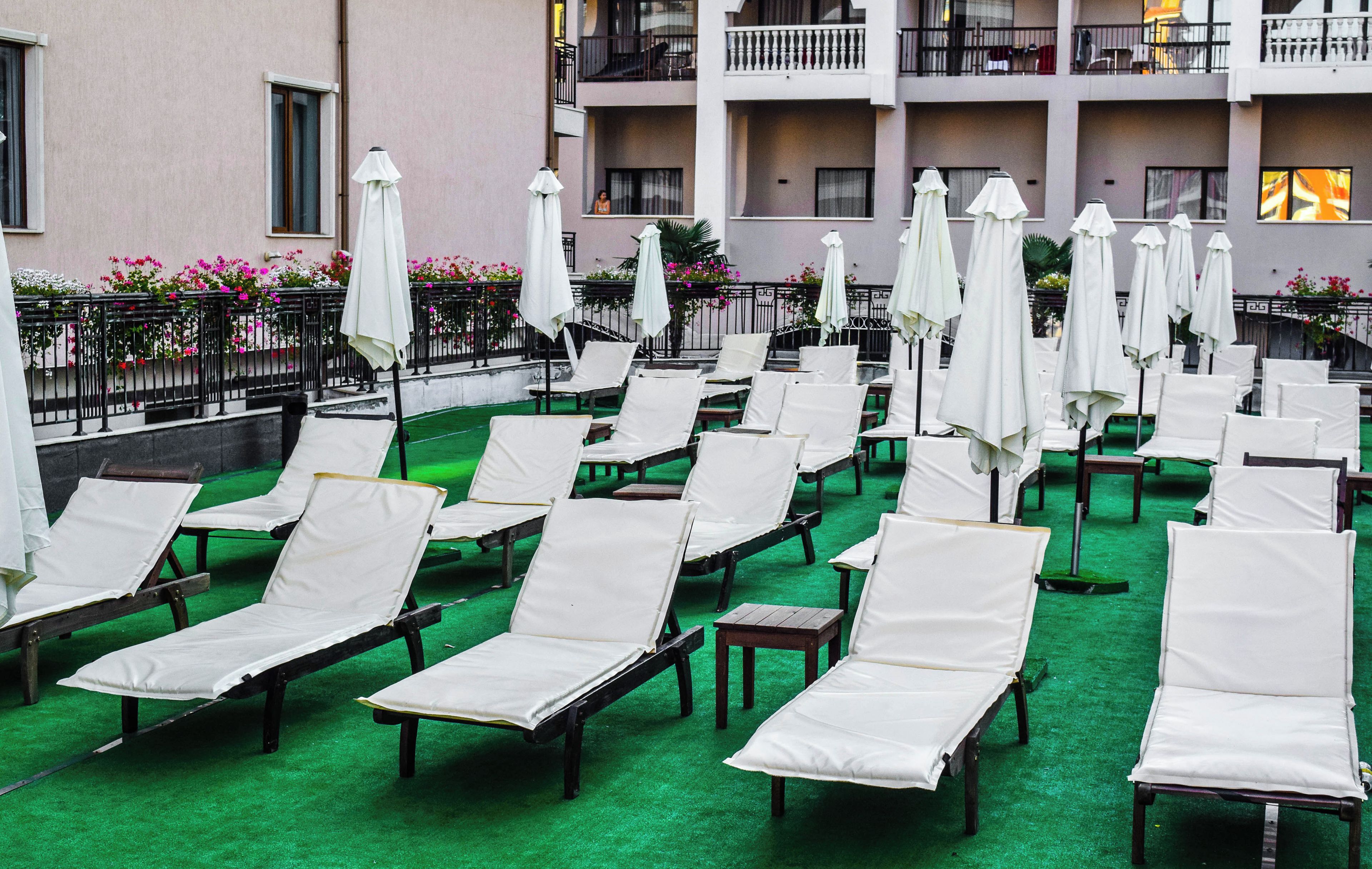 Eine Dachterrasse mit Reihen weißer Liegestühle und passenden Sonnenschirmen, einem grünen Teppich und einem Gebäude mit Balkonen im Hintergrund.