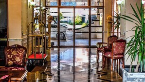Un vestíbulo con suelo a cuadros, sillas ornamentadas, una jaula de pájaros y un ventanal con vistas a un coche y follaje exterior.