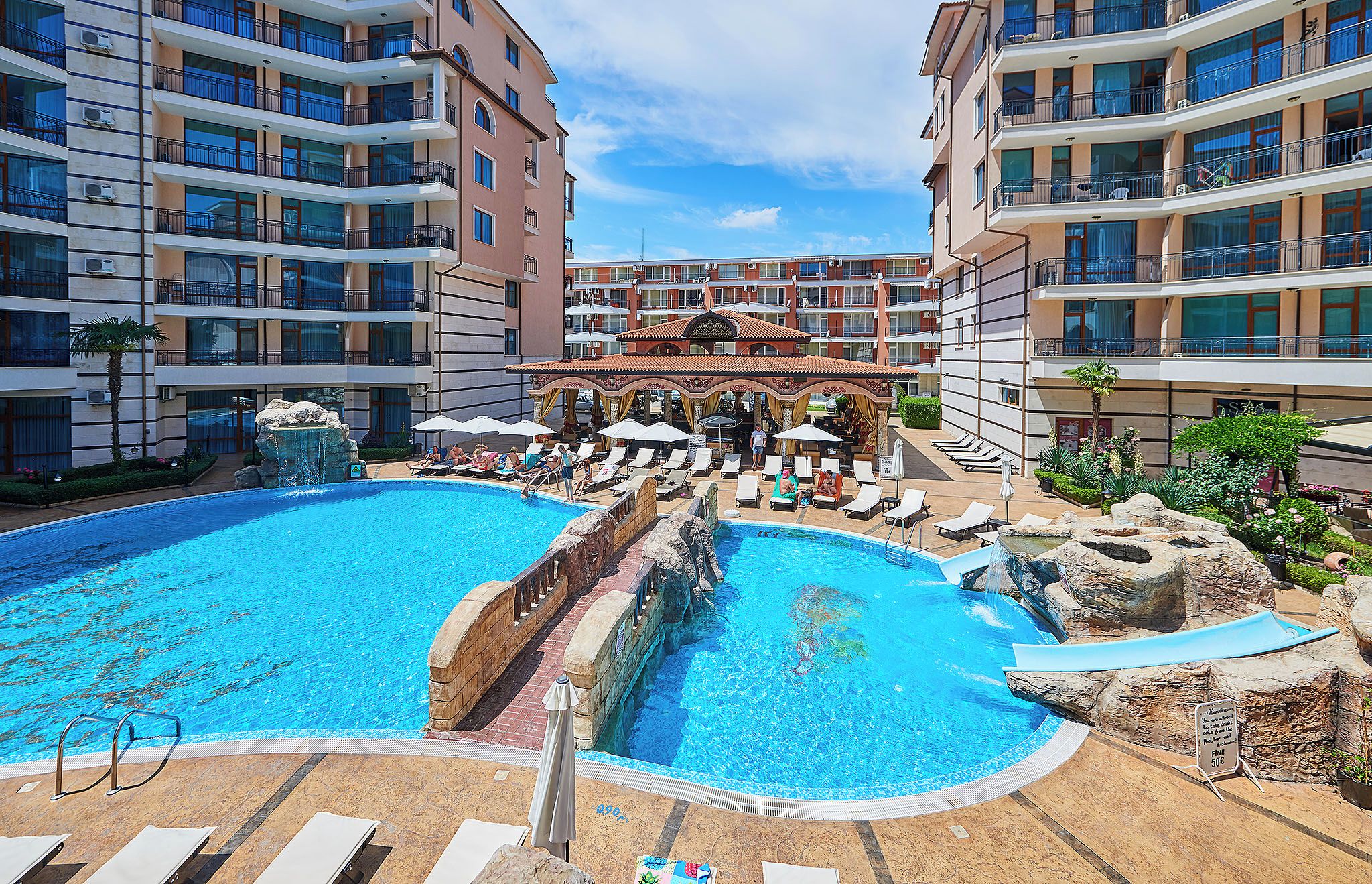 A multi-level hotel with a central pool area featuring a water slide, lounge chairs, and umbrellas.