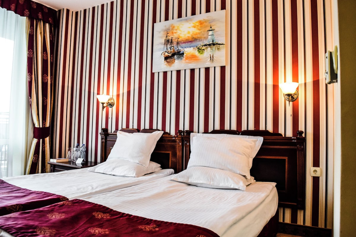 A hotel room with striped wallpaper, two beds with white linens, a painting of a sunset scene, and two wall-mounted light fixtures.