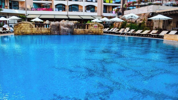 Una piscina con sillas de descanso y sombrillas, un elemento decorativo de cascada, y un edificio con balcones al fondo.