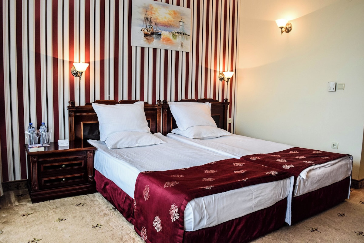 A double bed with a red bedspread, two bedside tables with lamps, a striped wallpaper background, and a painting above the bed.