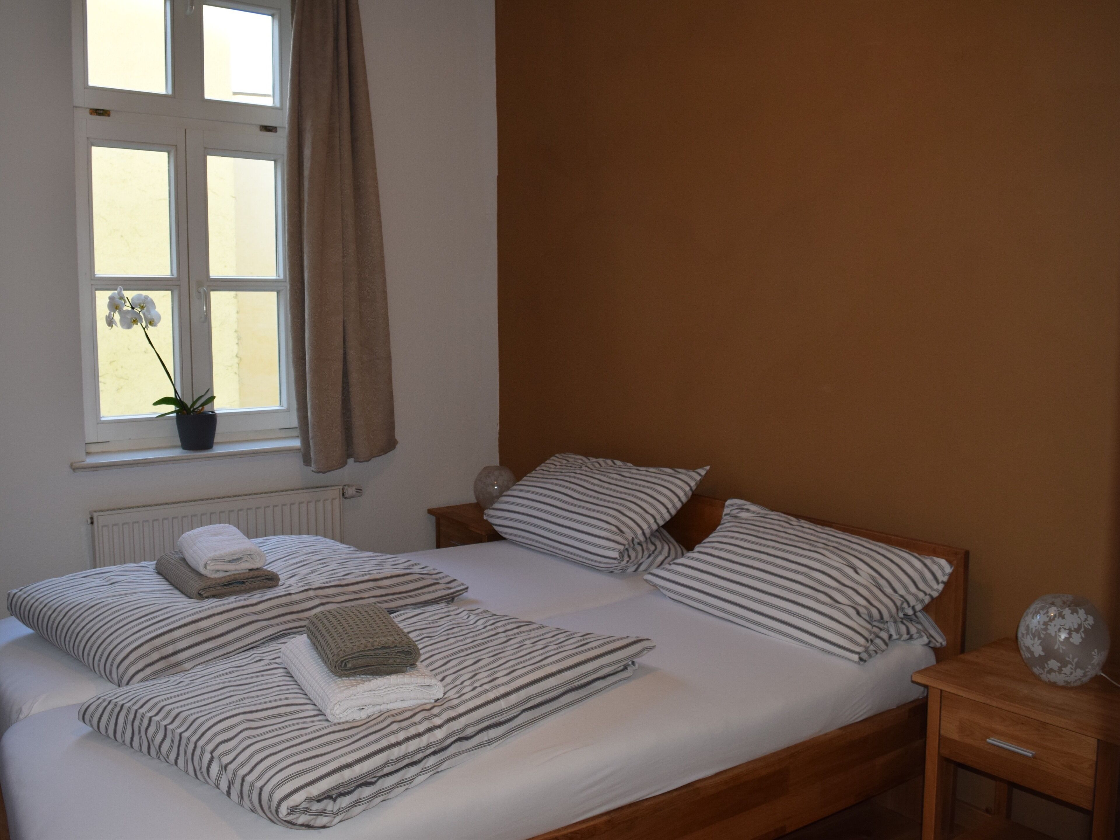 A neatly made bed with striped bedding and pillows, a window with curtains, a potted plant, and a bedside table with a lamp.