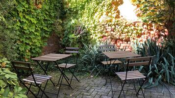 Un patio con mesas y sillas de madera, rodeado por un muro de ladrillo y abundante vegetación.