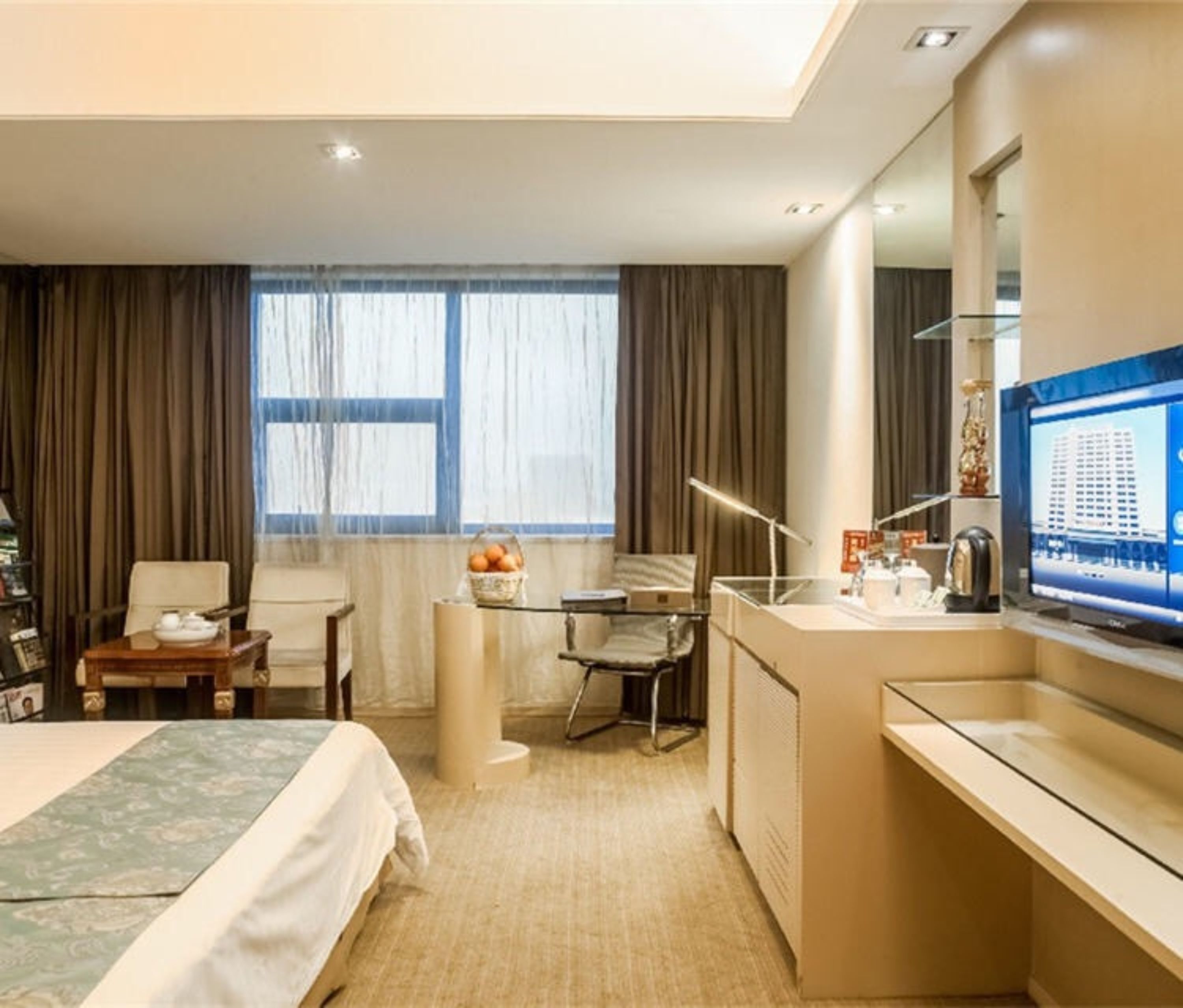 A hotel room with a bed, a desk with a computer, a chair, a small table with a basket of fruit, and a large window with curtains.