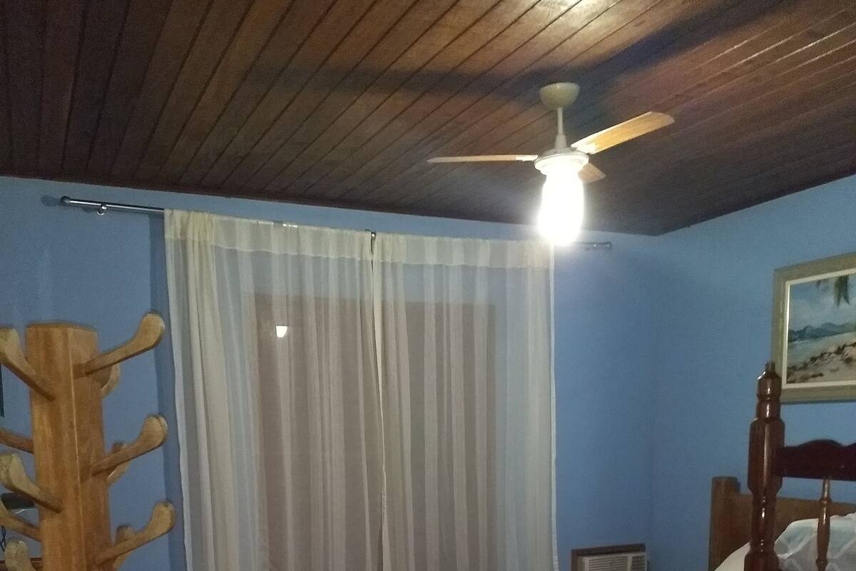 Une chambre avec deux lits en bois, un ventilateur de plafond, un tapis rouge à motifs et un rideau blanc.