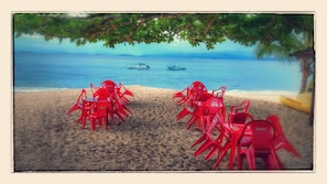 A beach with red plastic chairs arranged in rows, a sandy shore, and a calm sea with boats in the distance.