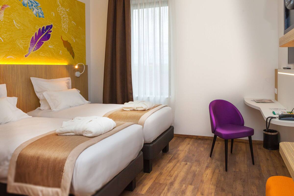 A hotel room with two beds, a desk, a purple chair, and a wooden floor.