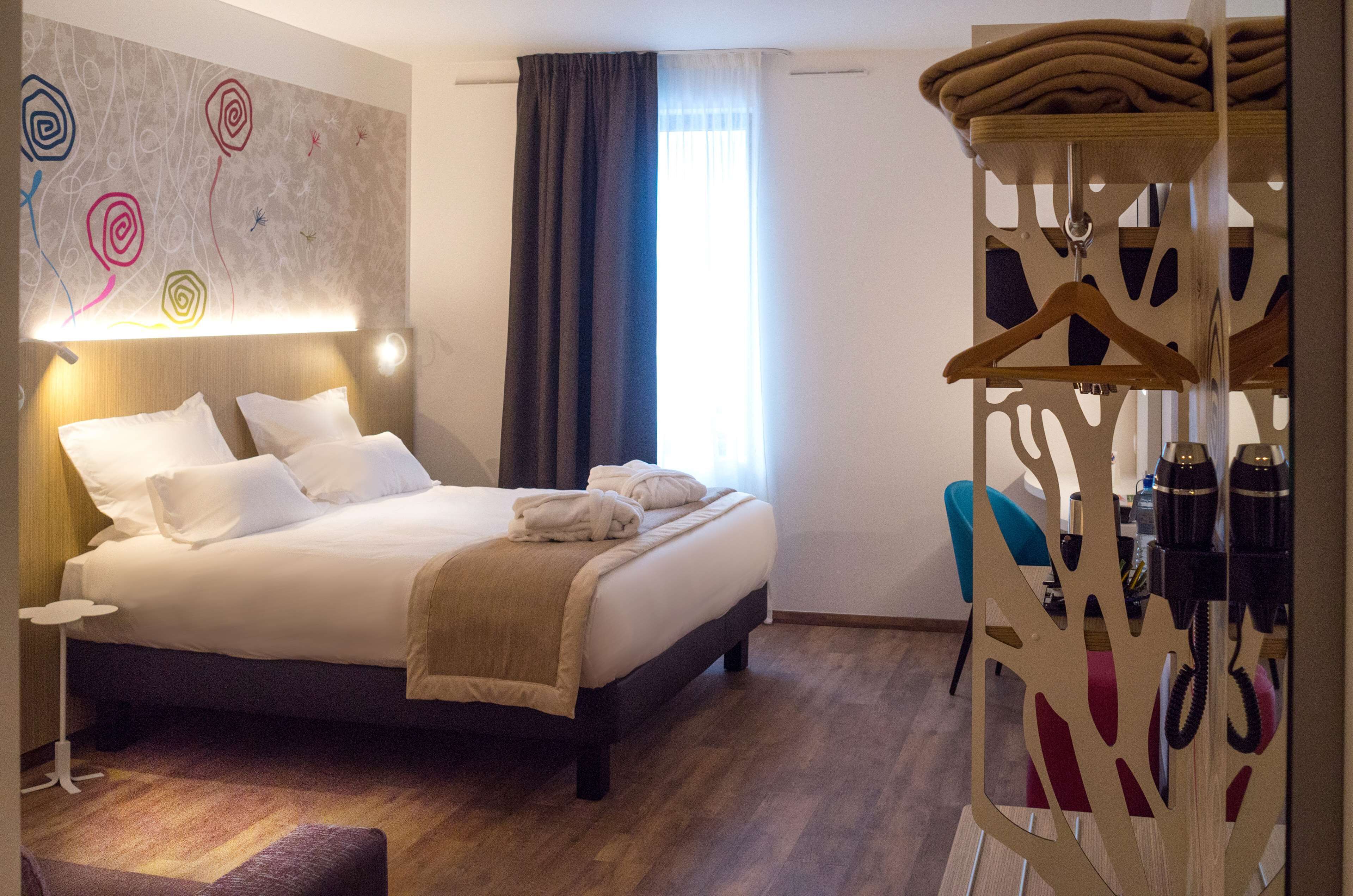 Ein Hotelzimmer mit einem Bett, einem Nachttisch, einer Wand mit abstrakter Kunst, einem Fenster mit Vorhängen und einem Holz-Kleiderständer mit Kleiderbügeln.