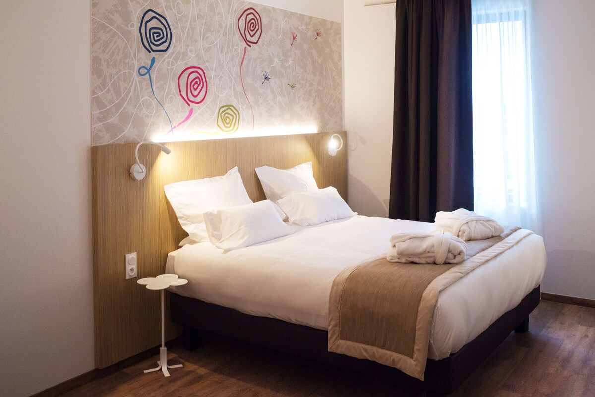 Uma cama bem arrumada com lençóis brancos e um cobertor bege, uma mesa de cabeceira com abajur, e uma parede com arte abstrata.