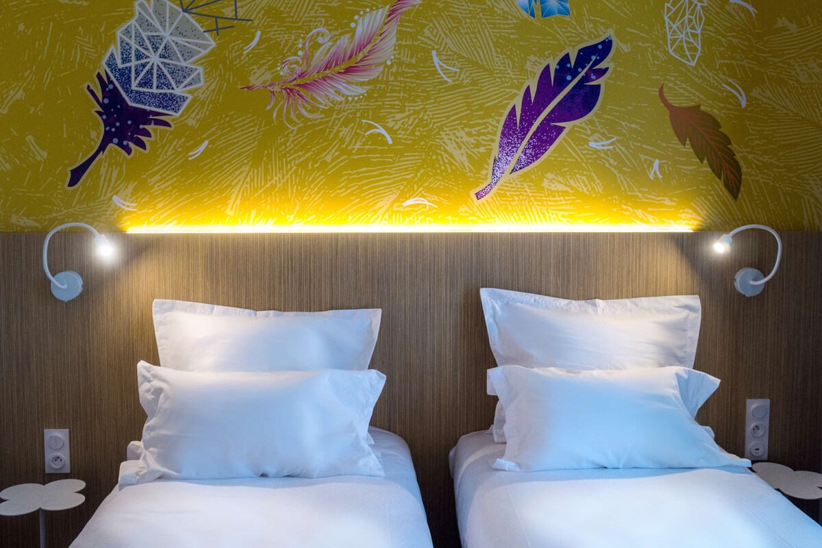 Quarto de hotel com duas camas, uma cabeceira de madeira e um mural com desenhos geométricos e de penas no teto.