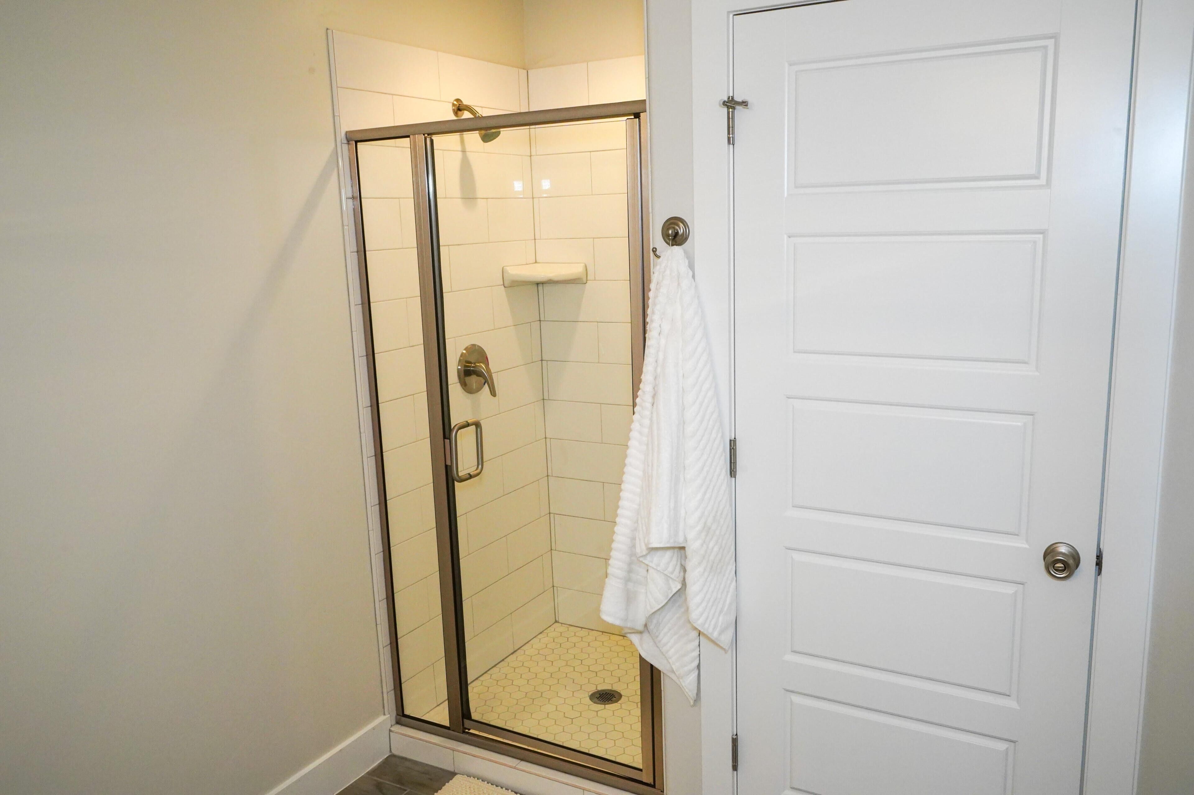 A glass-enclosed shower with a white towel hanging on the door, a white door with a silver handle, and a tiled floor.