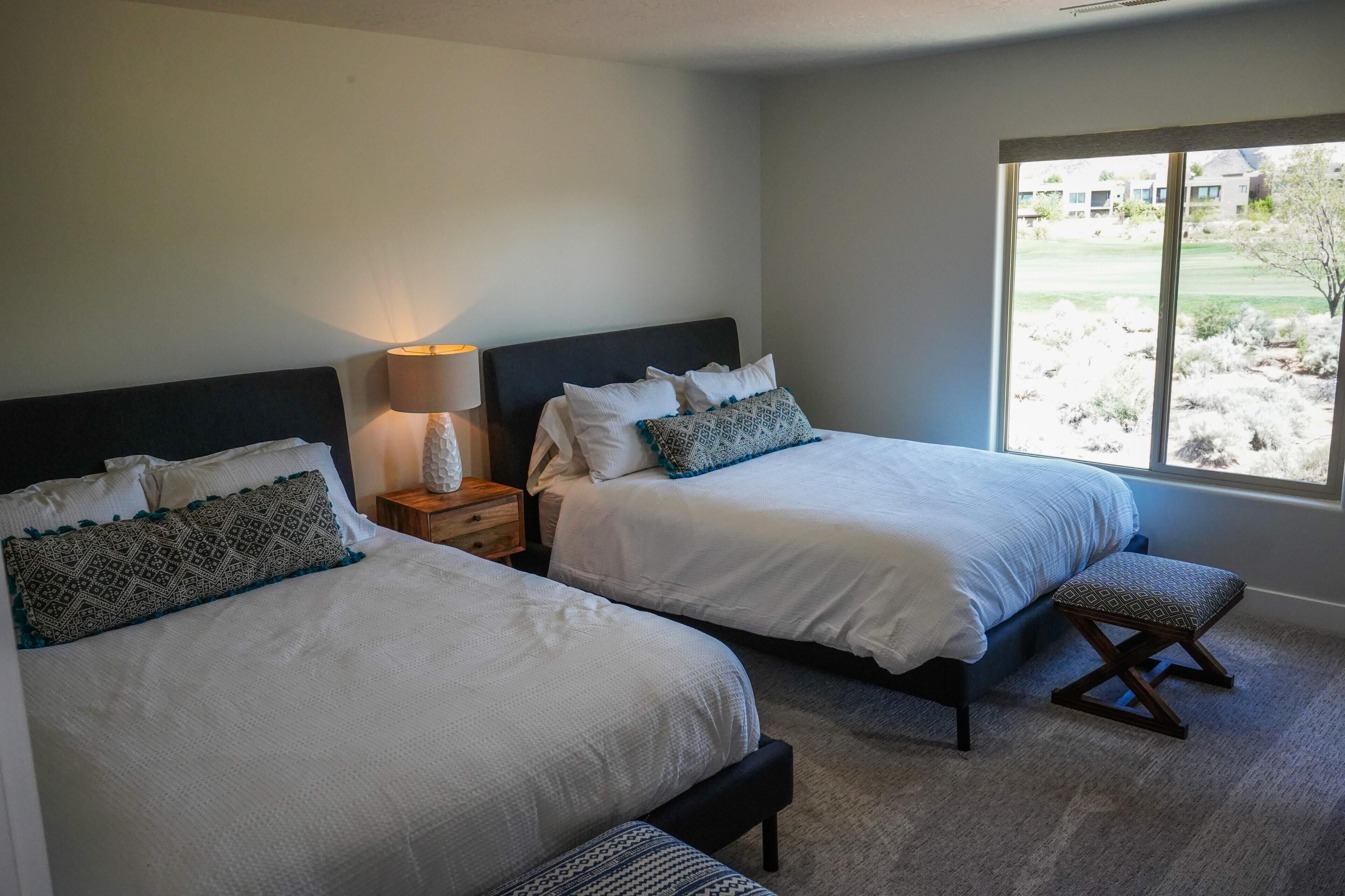 A bedroom with two beds, a nightstand, a lamp, and a window with a view of a landscaped area.