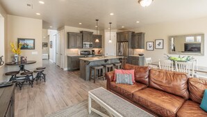 A spacious living area with a brown leather sofa, a coffee table, and a dining table with bar stools. The kitchen features dark wooden cabinets, stainless steel appliances, and pendant lighting. - Suite Escapes (St. George)