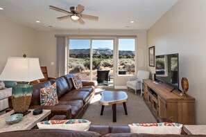 A living room with a leather couch, a coffee table, a TV, and a view of a desert landscape through large windows.