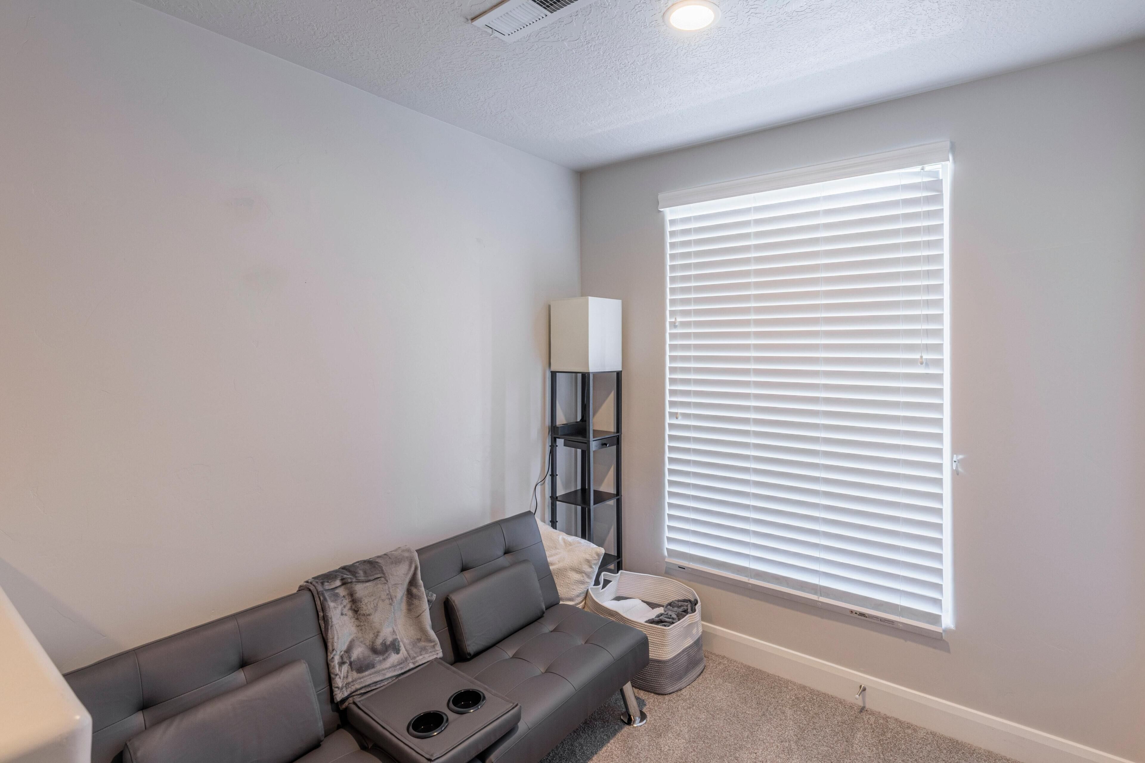 A modern living room with a grey sofa, a black shelving unit, a basket, and a large window with blinds.