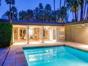 A modern house with a swimming pool, open doors leading to a living area, and tall palm trees in the background.