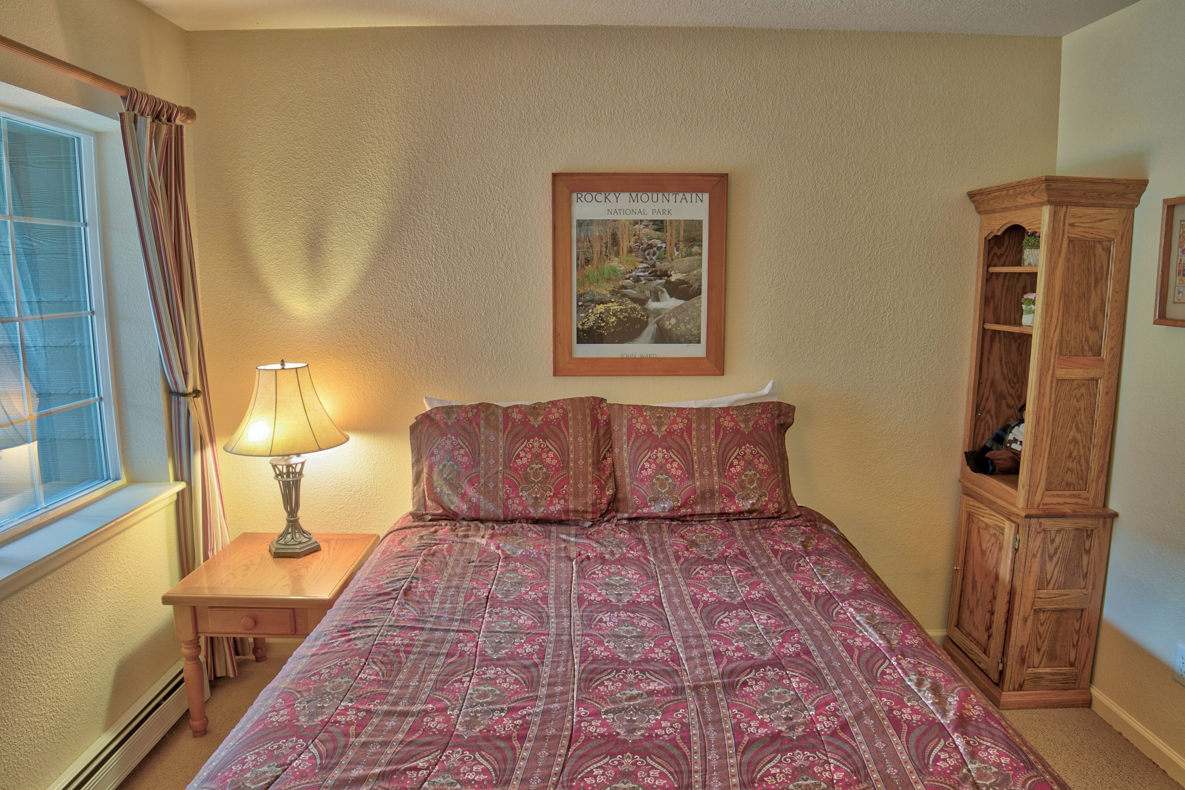 A bedroom with a bed, a nightstand with a lamp, a wooden cabinet, and a framed picture of Rocky Mountain National Park on the wall.