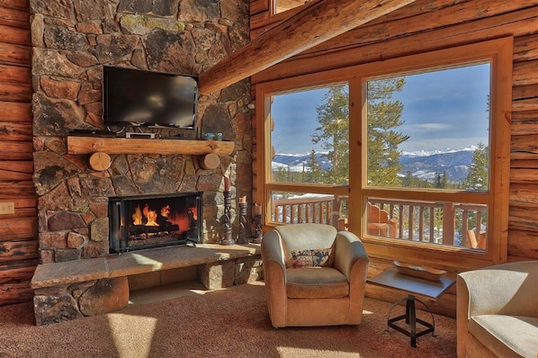 Interior acogedor de una cabaña de troncos con una chimenea de piedra, un televisor montado arriba y un ventanal con vistas a la montaña.