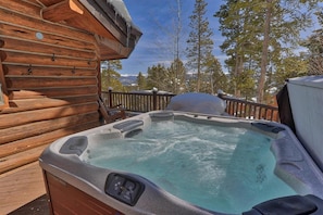 Una cabaña de madera con jacuzzi en una terraza, rodeada de árboles y montañas.