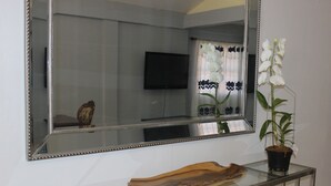 A living room with a large mirror, a flat-screen TV, a wooden decorative piece, and a potted orchid. - Ladyville 2Bed 2Bath (Ladyville)