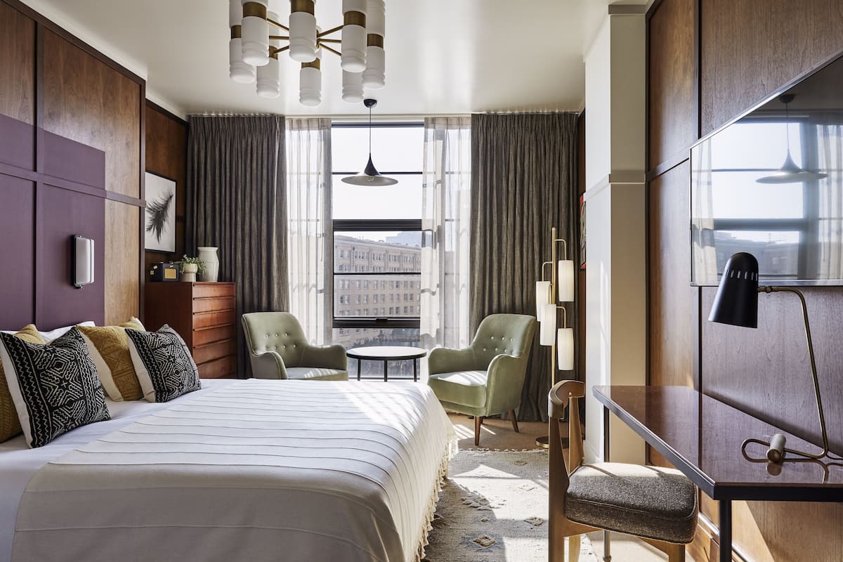A hotel room with a large bed, two armchairs, a small table, a desk, and a TV.