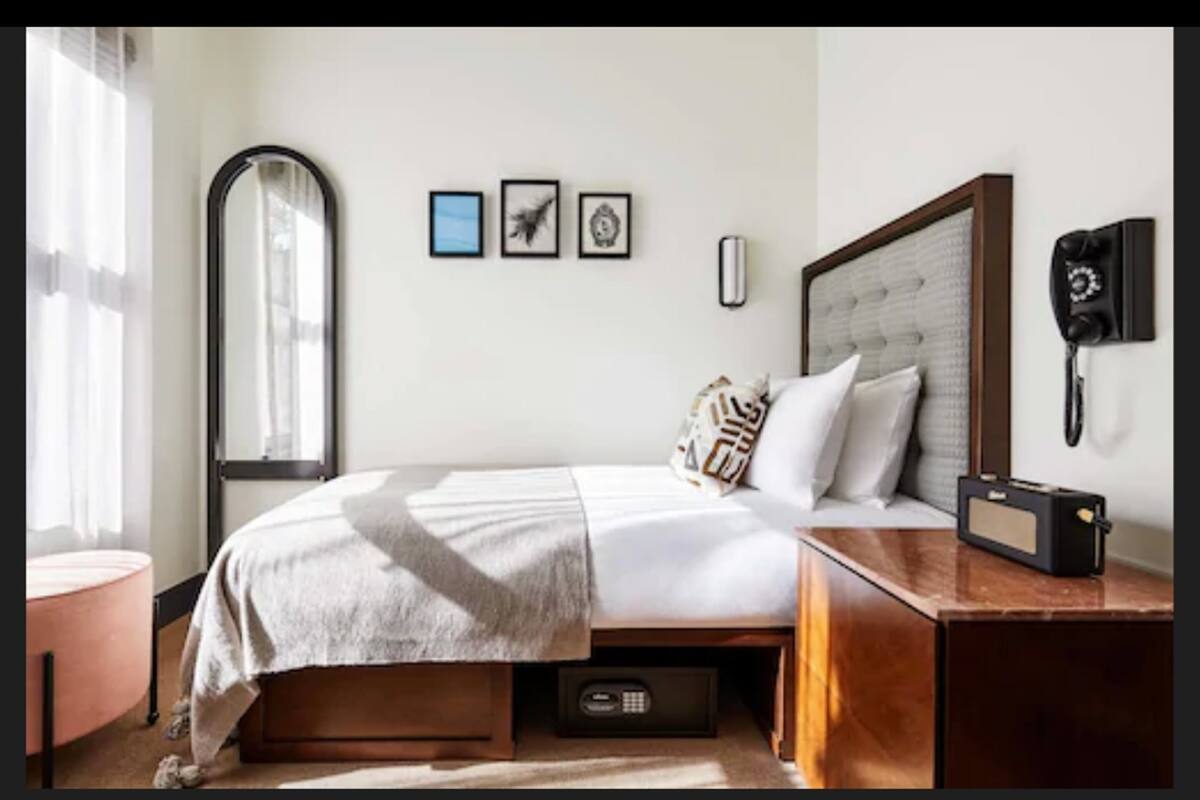 A neatly arranged hotel room with a bed, bedside table, and a mirror. There are three framed pictures on the wall and a telephone on the bedside table.