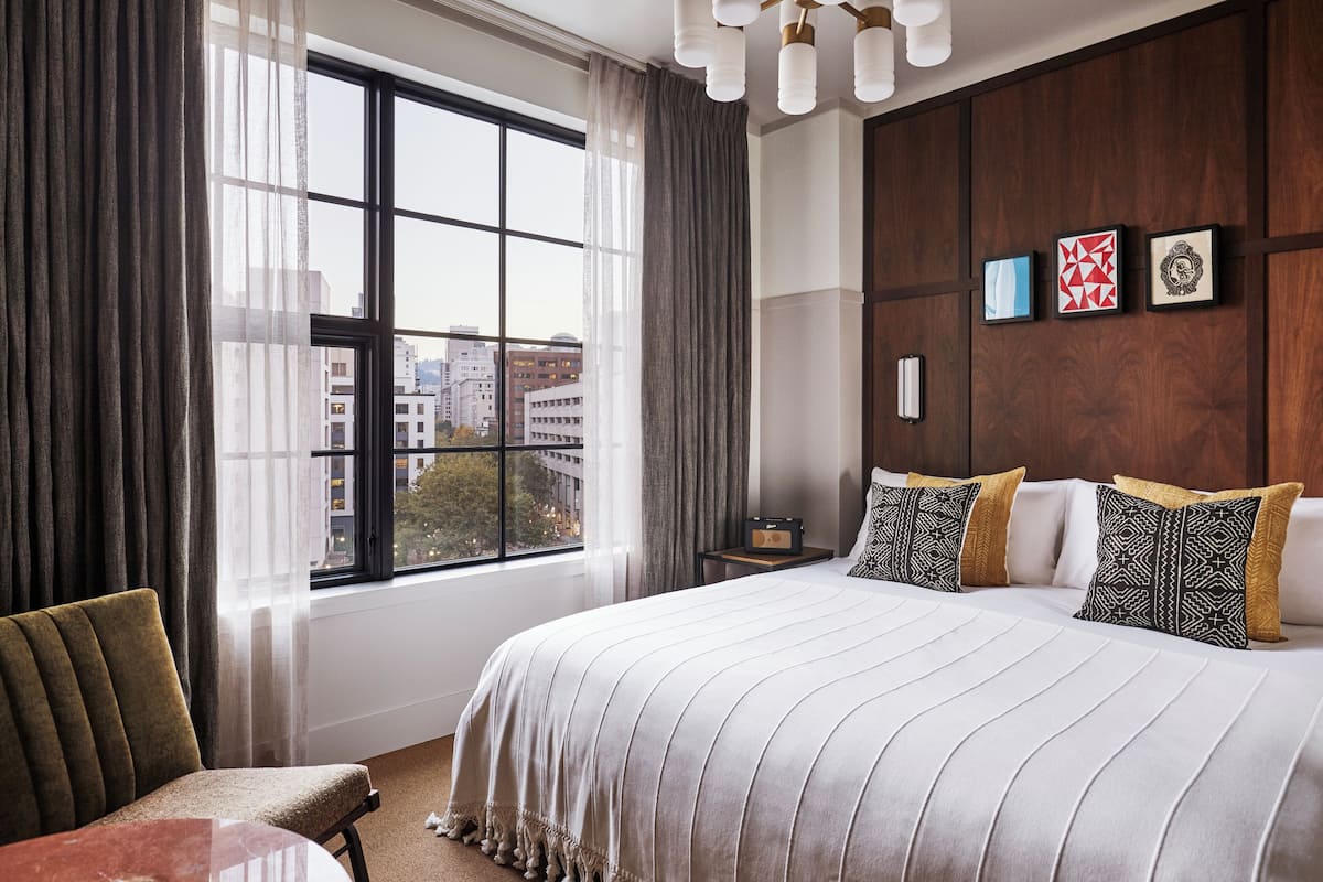 A hotel room with a large bed, a chair, a table, a window with curtains, and artwork on the wall.