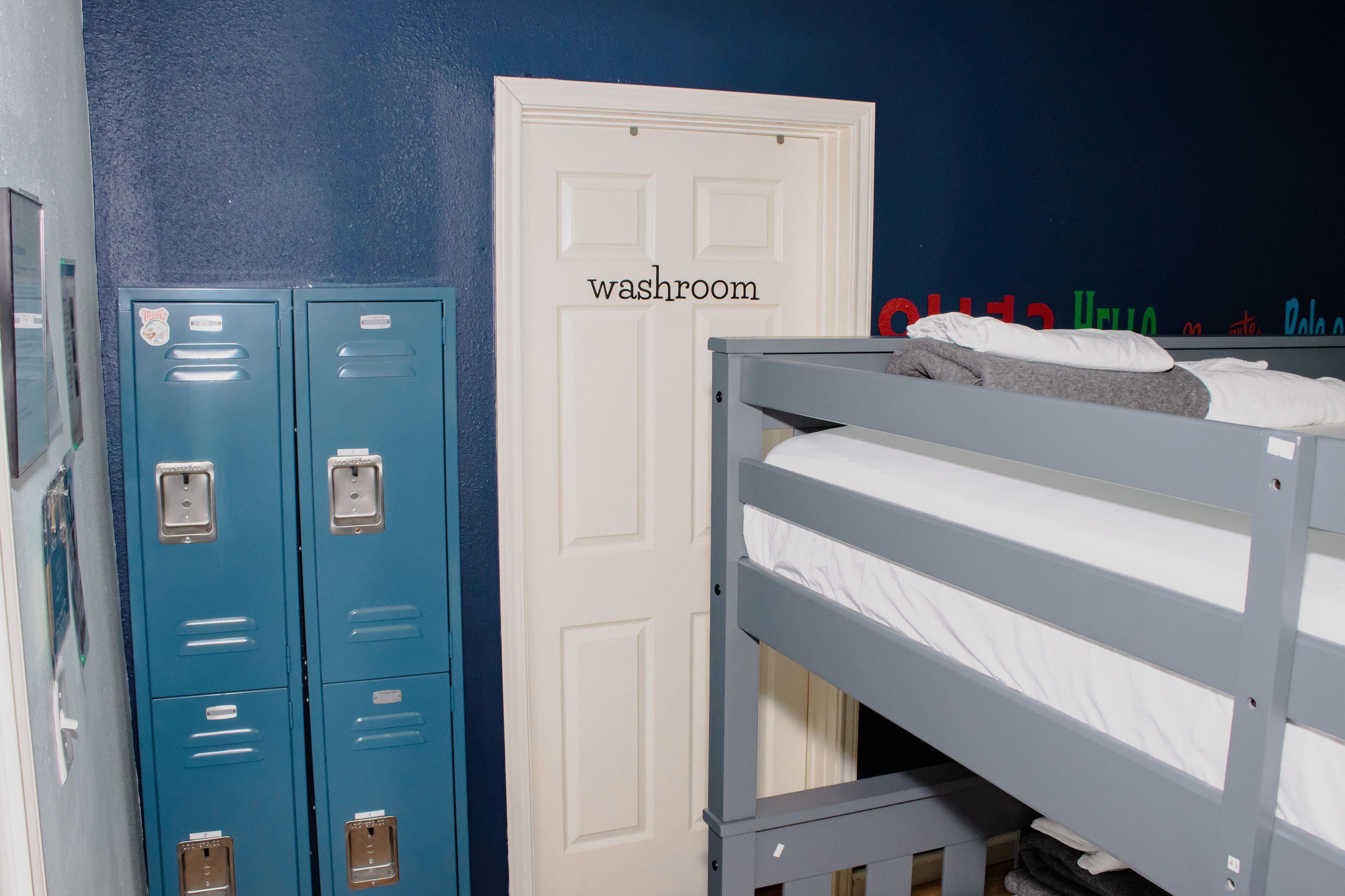 Una habitación con literas, una puerta blanca identificada como 'lavabo' y taquillas azules.