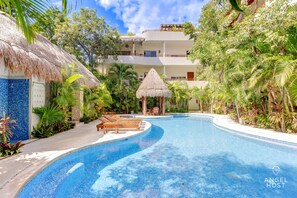 A swimming pool with a curved design, surrounded by lush greenery and a thatched-roof structure. - Condo Complex with an Alluring Pool & Tropical Vibes by Stella Rentals (Tulum)