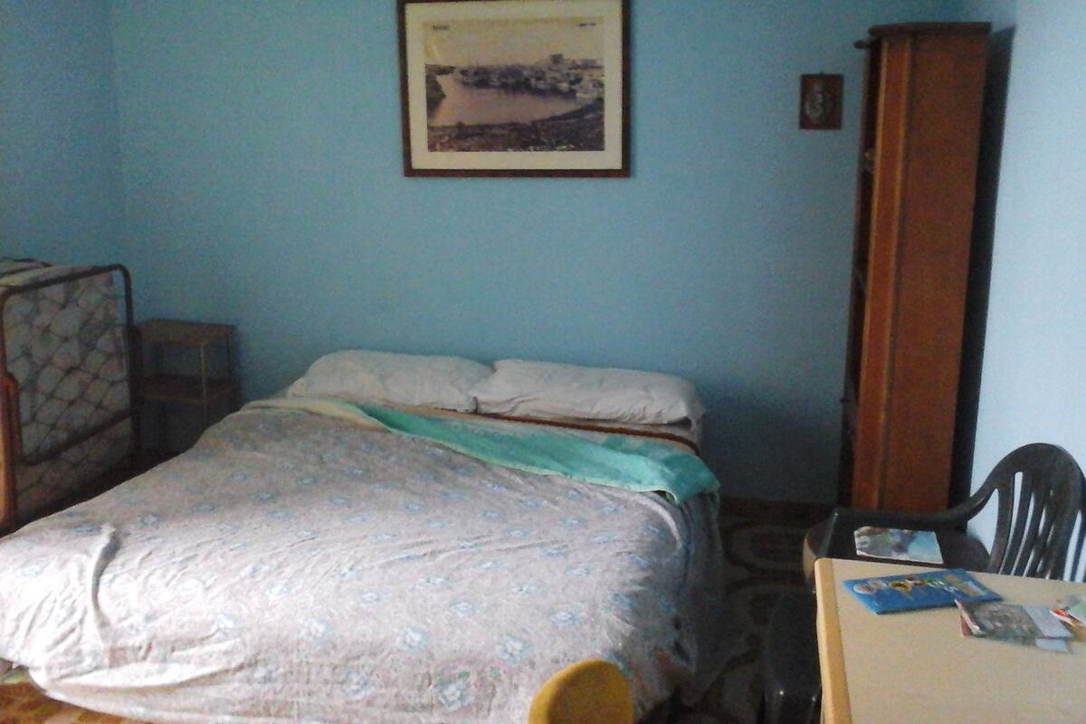 Ein kleiner, schlicht eingerichteter Raum mit einem Bett, einem Sessel, einem Tisch und einem hohen Schrank.