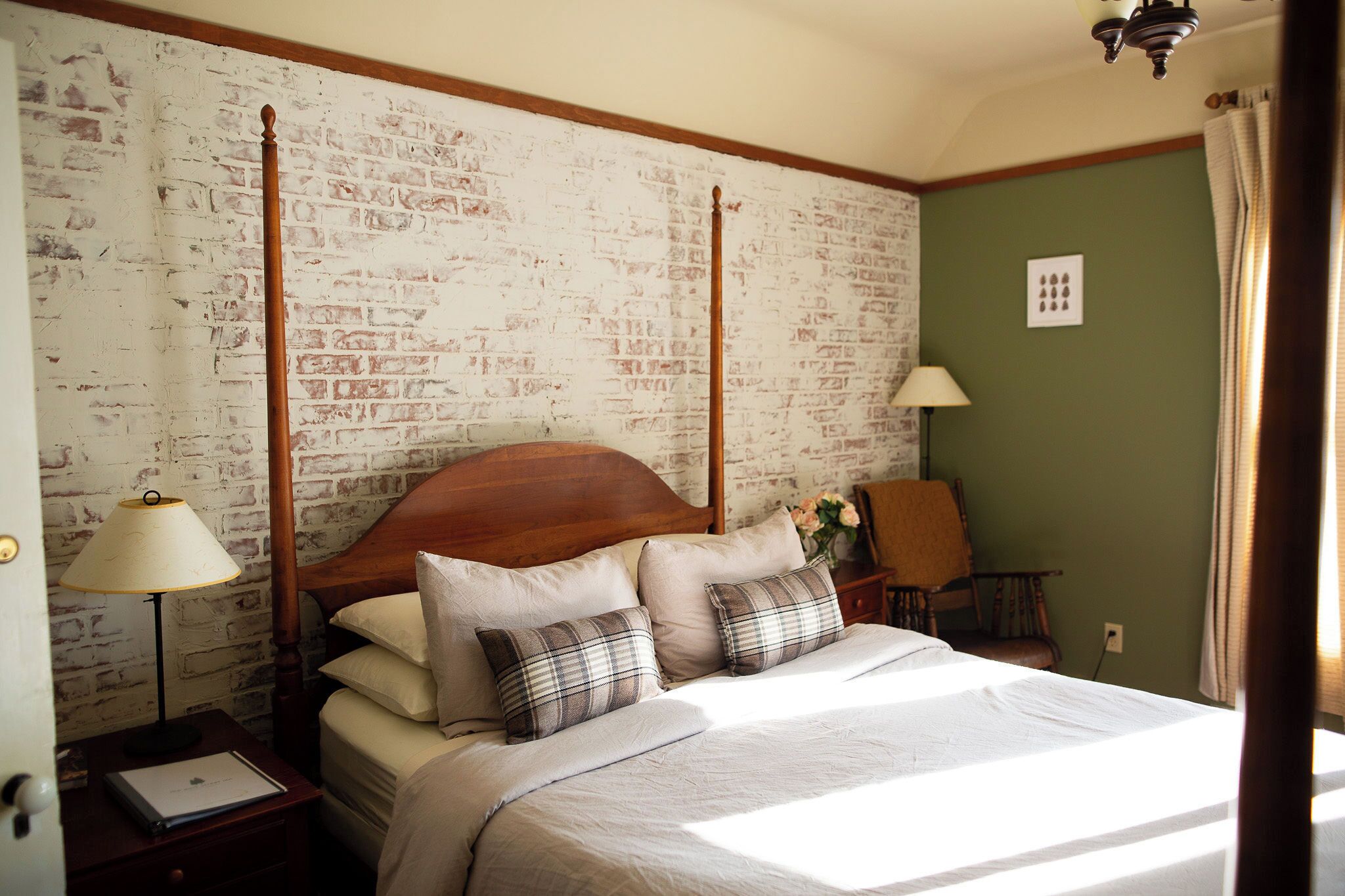 A neatly made bed with a wooden headboard, a nightstand with a lamp, and a chair in a room with brick-patterned wallpaper and green walls.