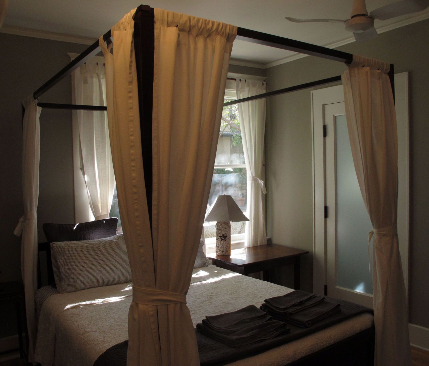 A four-poster bed with curtains, a bedside table with a lamp, and a view of the outdoors through a window.