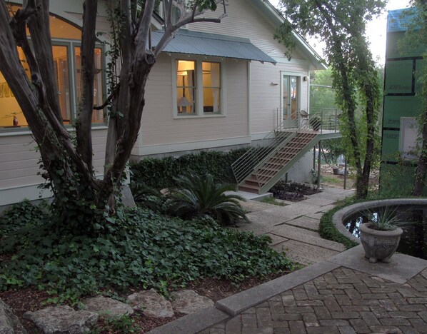 A house with a staircase leading to a balcony, surrounded by greenery and a small water feature.