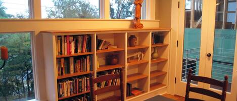 Une pièce avec une grande fenêtre, une bibliothèque remplie de livres, une table en bois avec un plateau et une chaise.