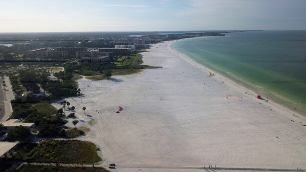 Vista aérea de una playa con cielo despejado, orilla arenosa y edificios al fondo.