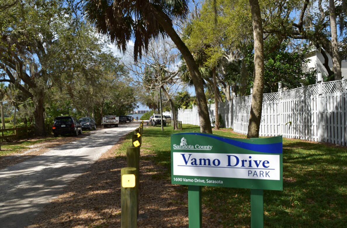 A park entrance sign for Vamo Drive Park in Sarasota County, with a road leading into the park and vehicles parked along the side.