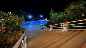 Vista nocturna de un área de piscina con una escalera de madera que baja a la piscina, rodeada de exuberante vegetación e iluminación decorativa.