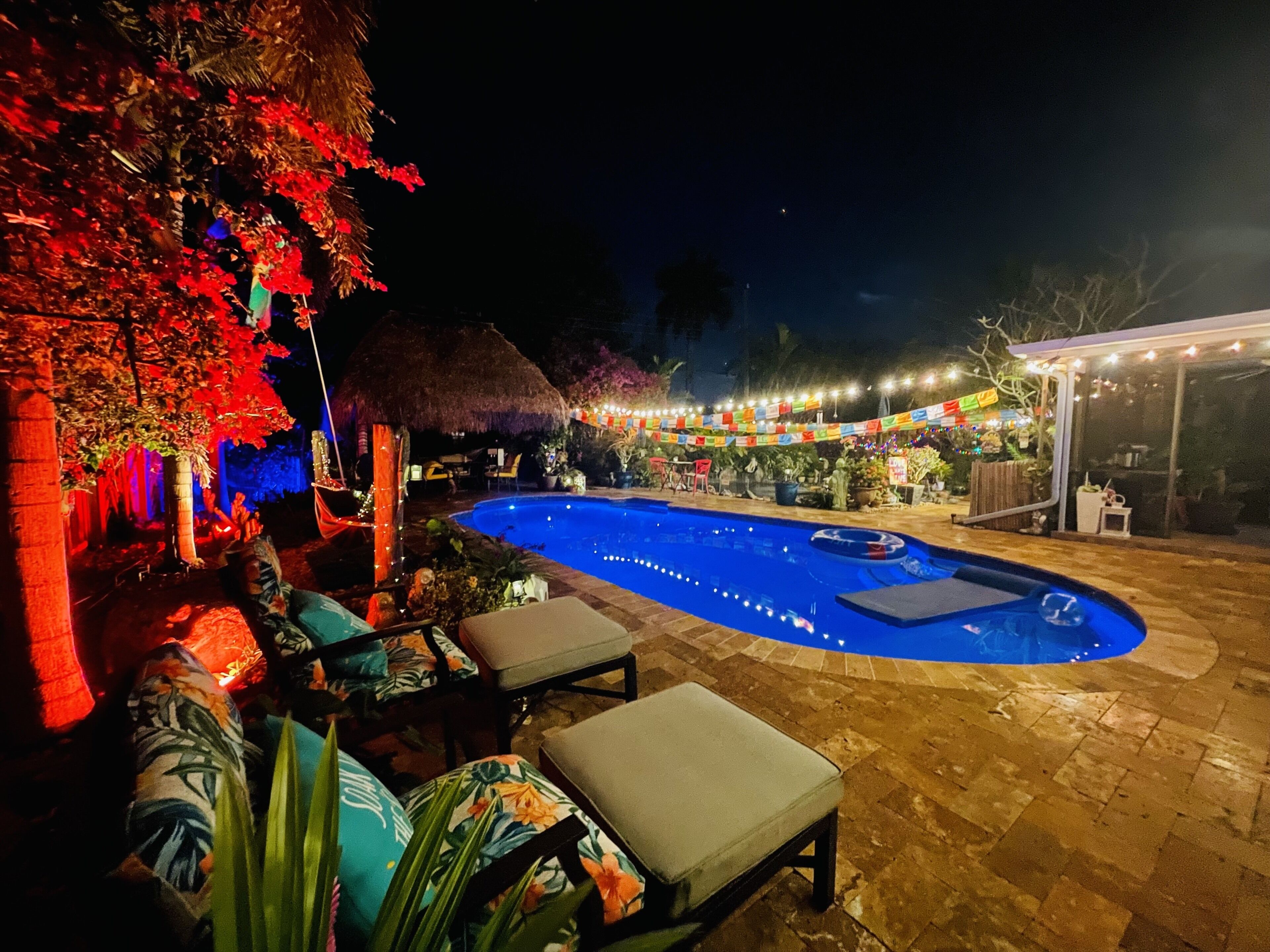 A nighttime outdoor pool area with a thatched-roof structure, lounge chairs, and a covered seating area.