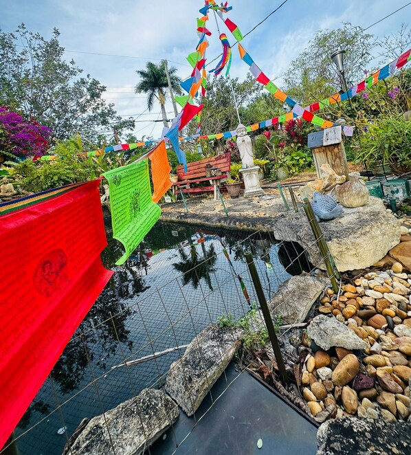 A serene outdoor area with a small pond, colorful prayer flags, and a Buddha statue.