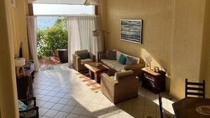 A living room with a sofa, armchairs, a coffee table, and a dining table. There is a window with curtains, a painting on the wall, and a view of the outdoors.