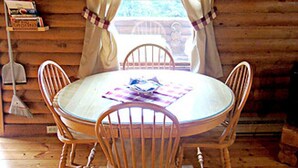 Un intérieur en bois avec une table ronde et quatre chaises, un lambrequin à carreaux au-dessus d’une fenêtre et un balai appuyé contre le mur.