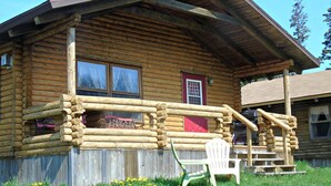 Une cabane en rondins avec un porche, une porte rouge et une chaise blanche à l’extérieur.