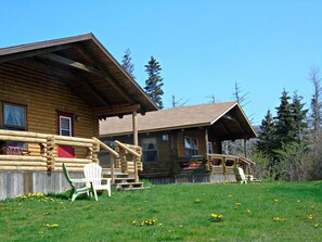 Une cabane en rondins dotée d’une véranda et d’un espace abrité, entourée d’un champ verdoyant et d’arbres.