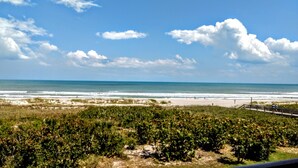 A beach view with a wooden boardwalk, grassy areas, and dense vegetation in the foreground.