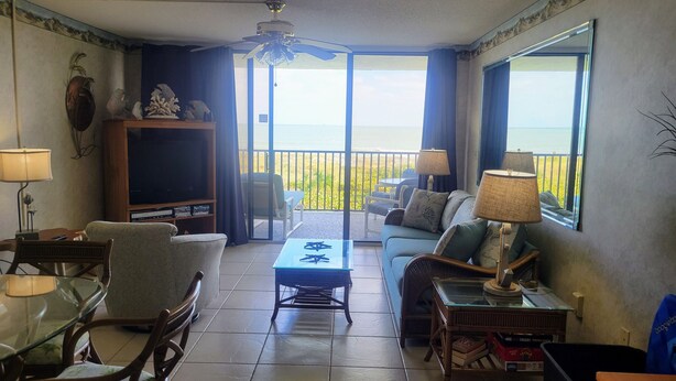 A living room with a glass table, chairs, a sofa, a ceiling fan, and a view of the ocean through large windows.