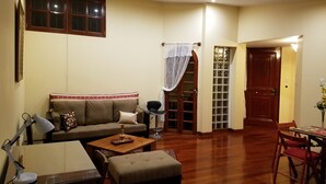 Una sala de estar con sofá, mesa de centro y reposapiés, una mesa de comedor con sillas, y una puerta que da a otra habitación.